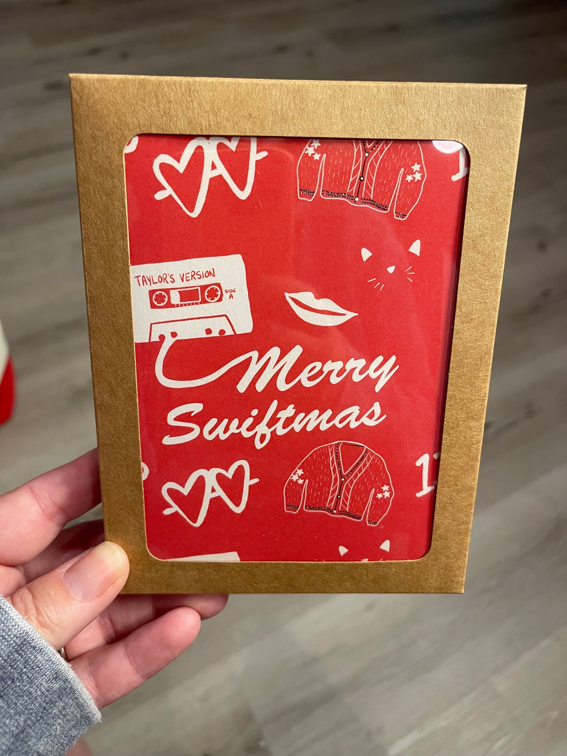 Greeting card that reads merry swiftmas on the front with taylor swift inspired icons on a red background. Shown in a boxed card set