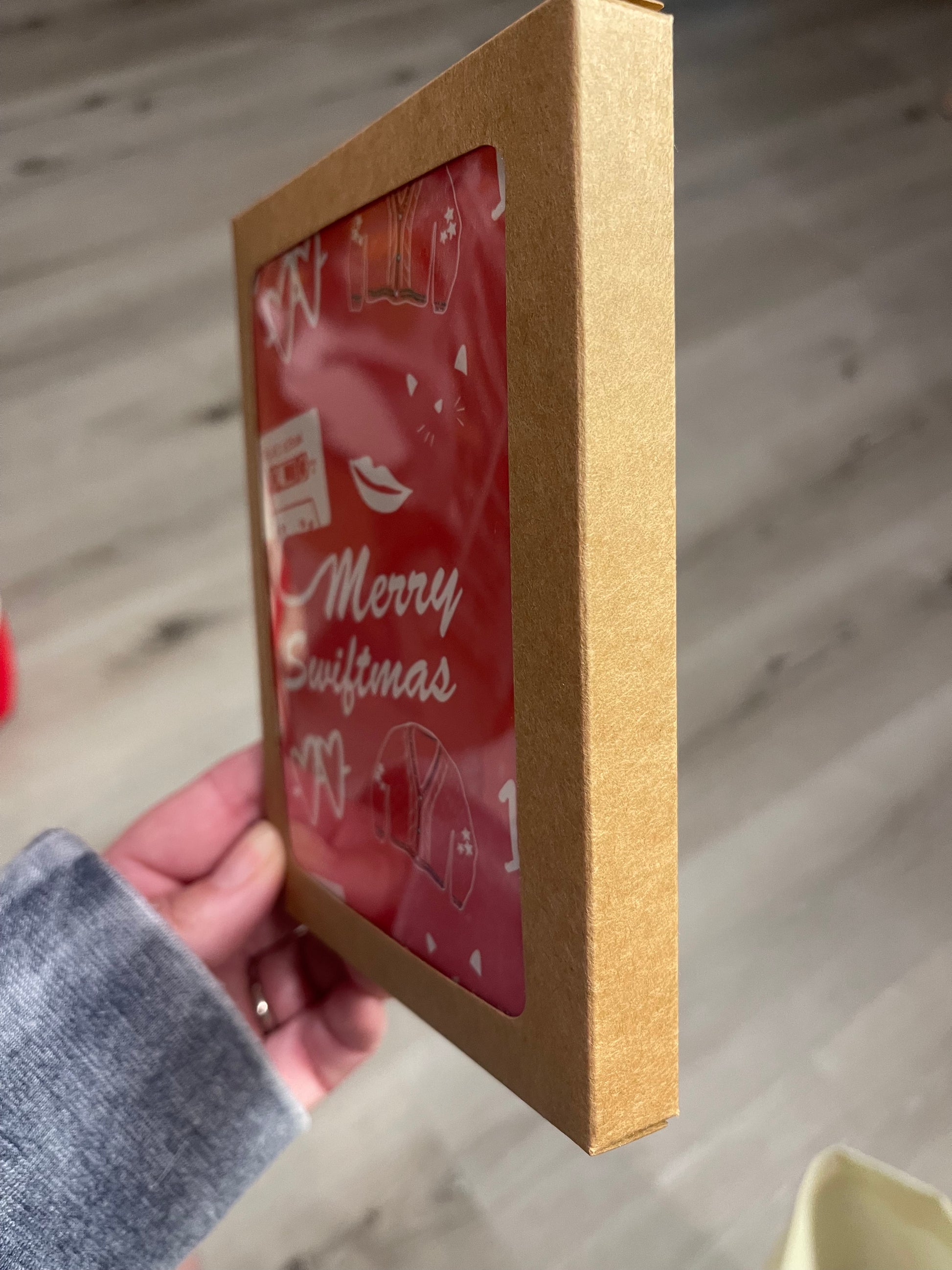 Greeting card that reads merry swiftmas on the front with taylor swift inspired icons on a red background. Shown in a boxed card set